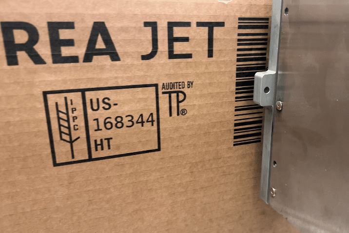 Głowica drukująca UP REA JET nanosi czarny nadruk z logo, kodem kreskowym oraz oznaczeniami certyfikacyjnymi na brązowym kartonie w przemysłowej linii produkcyjnej.