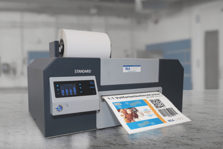 Przemysłowa drukarka REA LABEL ColorJet drukująca kolorową etykietę z kodem QR, grafiką i zmiennymi danymi, przeznaczoną do znakowania produktów i opakowań.