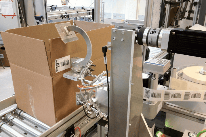 Automatyczny aplikator etykiet REA LABEL nakłada etykietę z kodem kreskowym i danymi logistycznymi na karton podczas procesu pakowania.