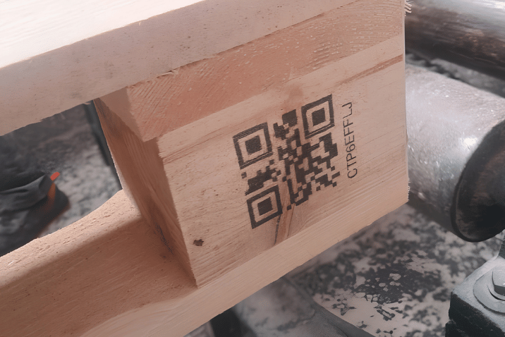 Zbliżenie na klocek palety drewnianej z nadrukowanym kodem QR i oznaczeniem alfanumerycznym.