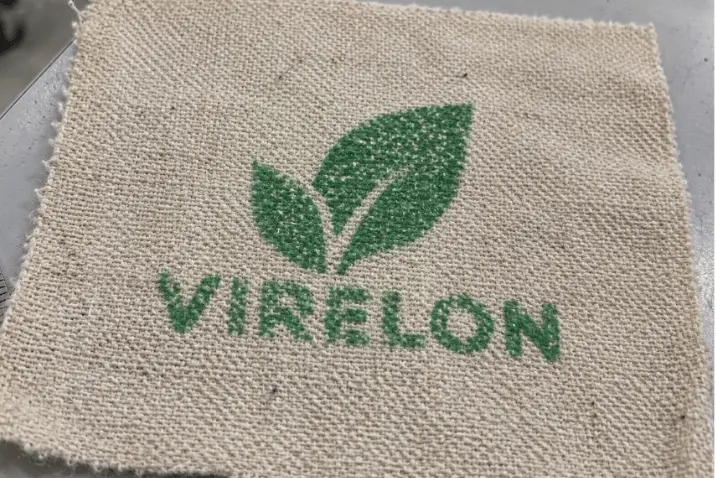 Zielony nadruk logo z motywem liści na jasnej tkaninie o widocznym splocie i naturalnej strukturze włókien.