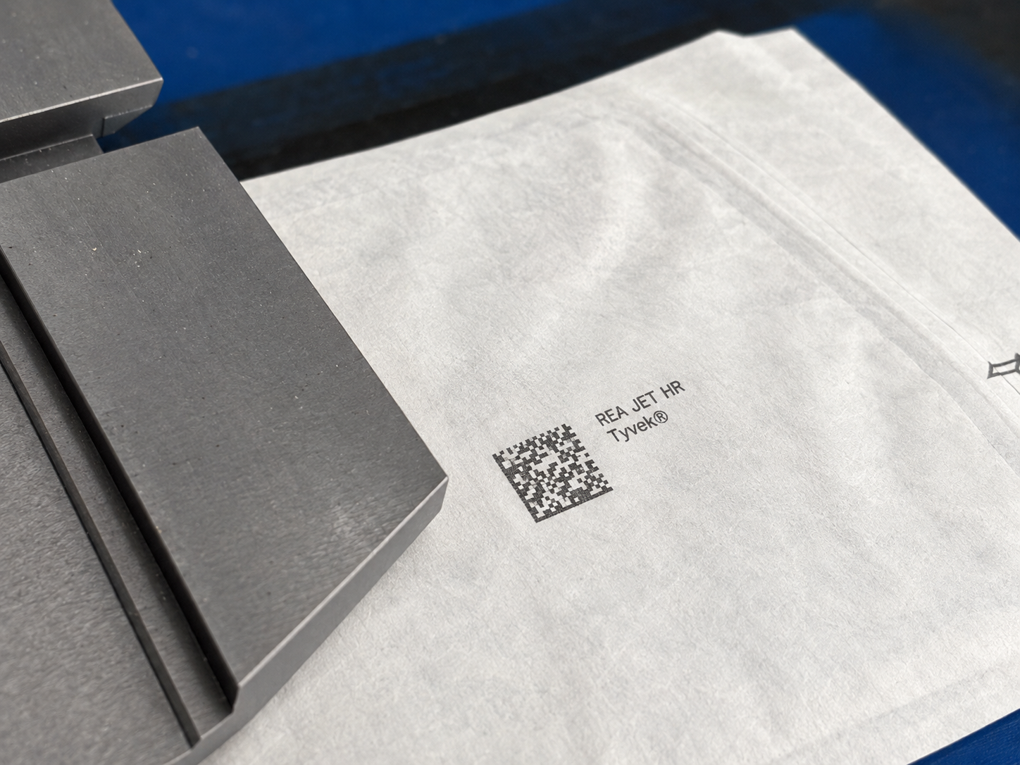 Zbliżenie na kod DataMatrix i oznaczenie REA JET HR nadrukowane na białym materiale Tyvek przy przemysłowej prowadnicy.